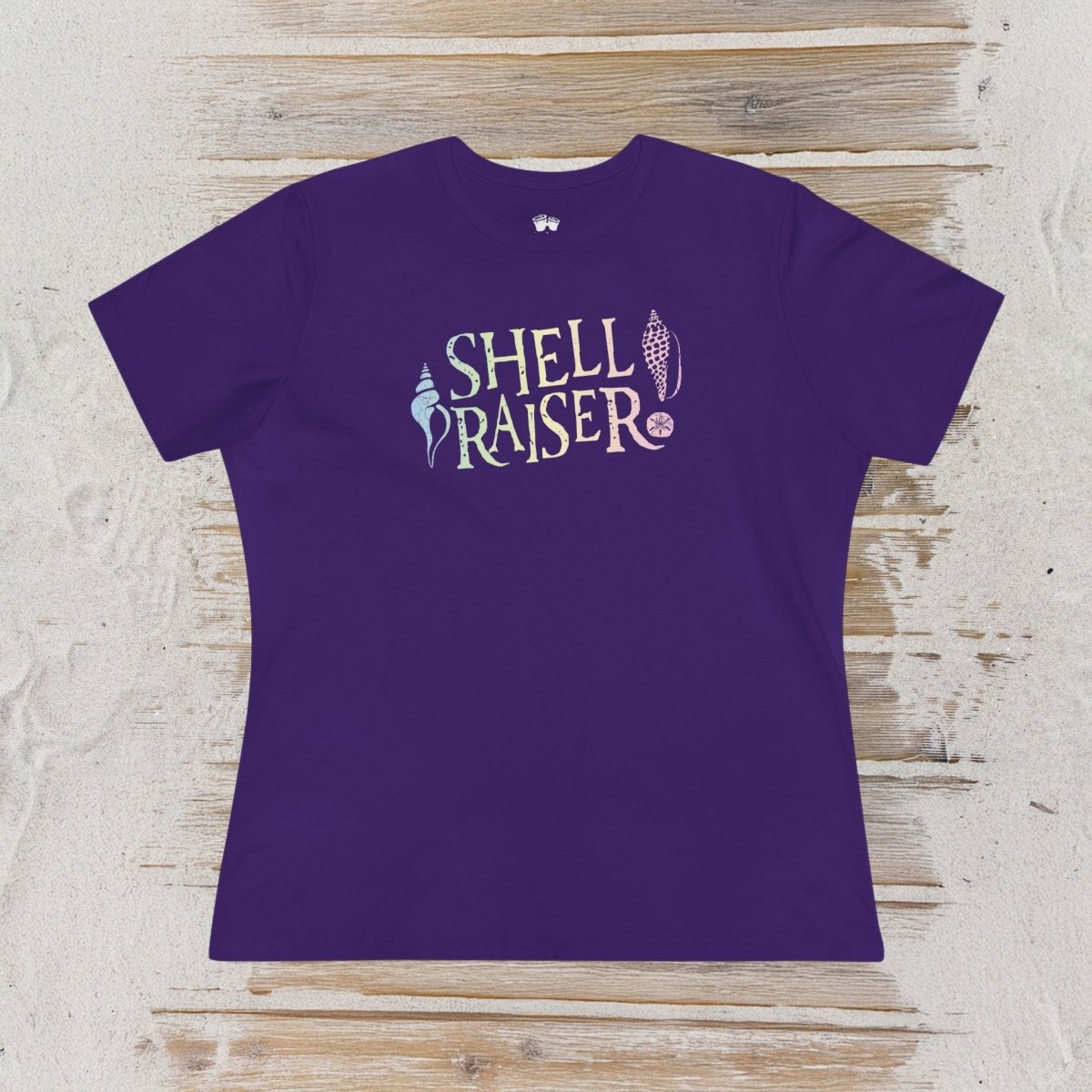 A purple short-sleeve t-shirt with a 'SHELL RAISER' graphic on the chest, laid flat on a light-colored wooden plank surface.