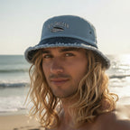 Surfer dude wearing a light blue bucket hat with a dark navy brim and 'OLD SHELTER' over a Junonia shell embroidered on the front.