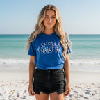 Beachwear model in a royal blue short-sleeve t-shirt that features a graphic print reading 'SHELL RAISER' in a stylized white and light blue paltel font, accompanied by illustrations of seashells