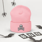 A light pink knit beanie with a folded cuff featuring a gray embroidered skull and crossbones. The beanie is displayed on a white surface decorated with Halloween props, including hanging spiders and 'BOO' letter cutouts.