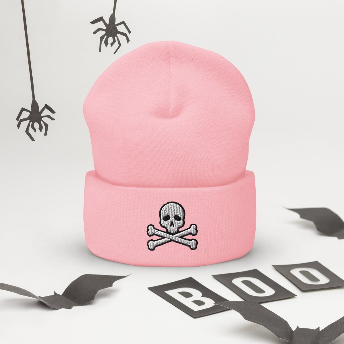 A light pink knit beanie with a folded cuff featuring a gray embroidered skull and crossbones. The beanie is displayed on a white surface decorated with Halloween props, including hanging spiders and 'BOO' letter cutouts.