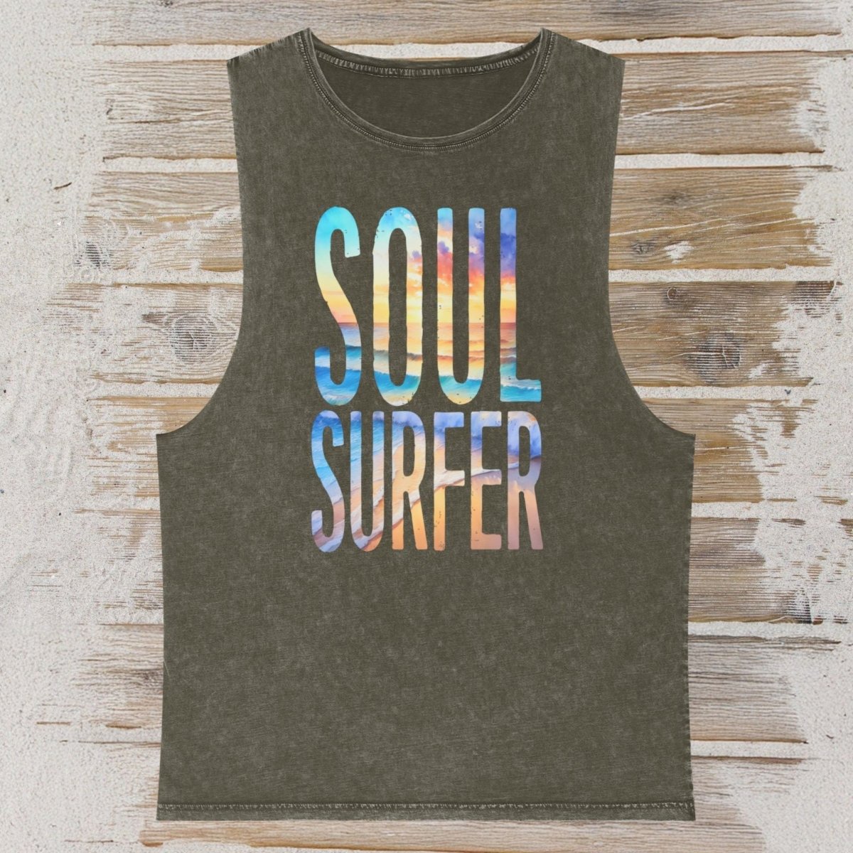 An olive green sleeveless tank top with 'SOUL SURFER' printed in a colorful beach scene font, laid flat on a rustic wood plank background.