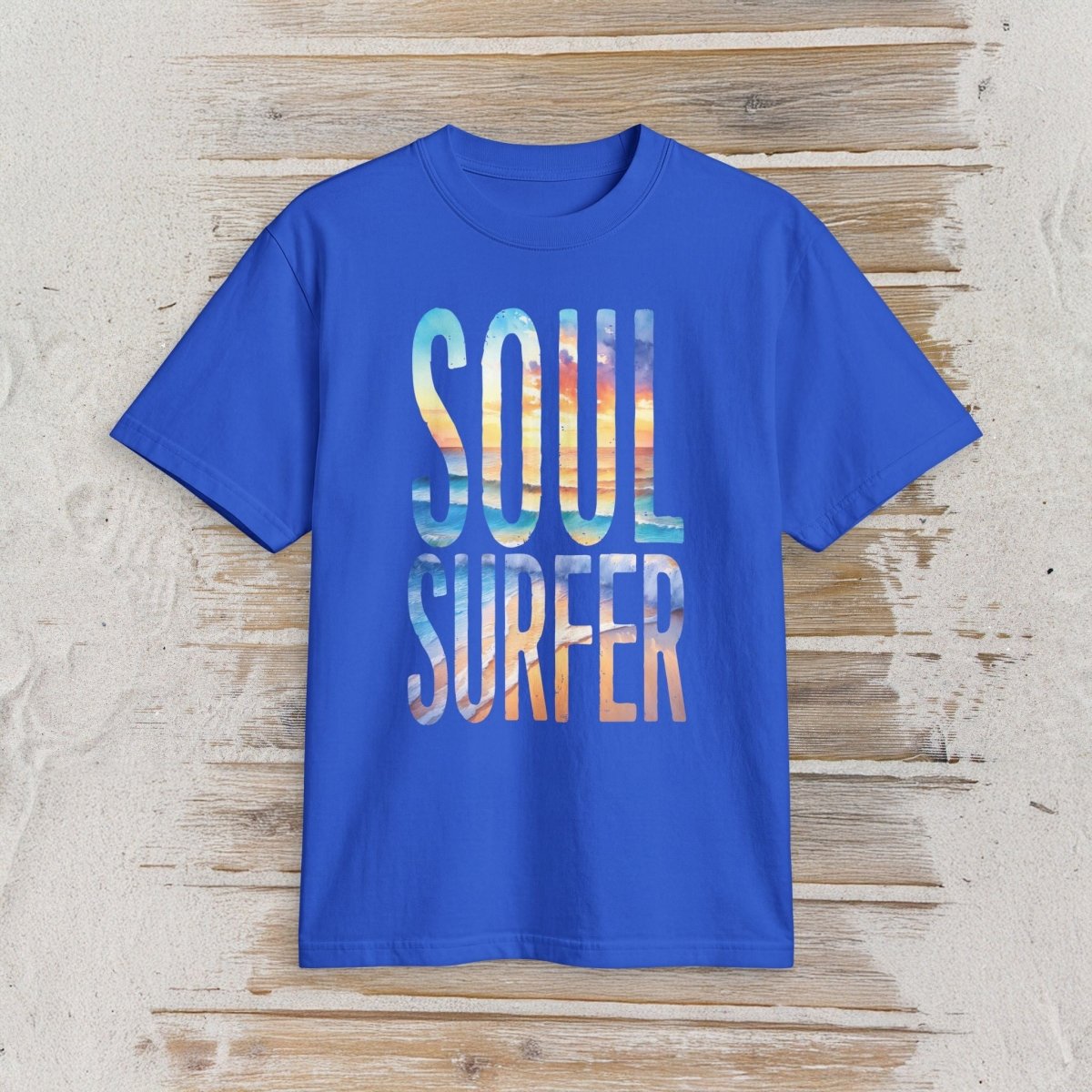A royal blue short-sleeve t-shirt with 'SOUL SURFER' printed in a colorful, textured beach scene font on the front, displayed flat against a rustic light wood background.