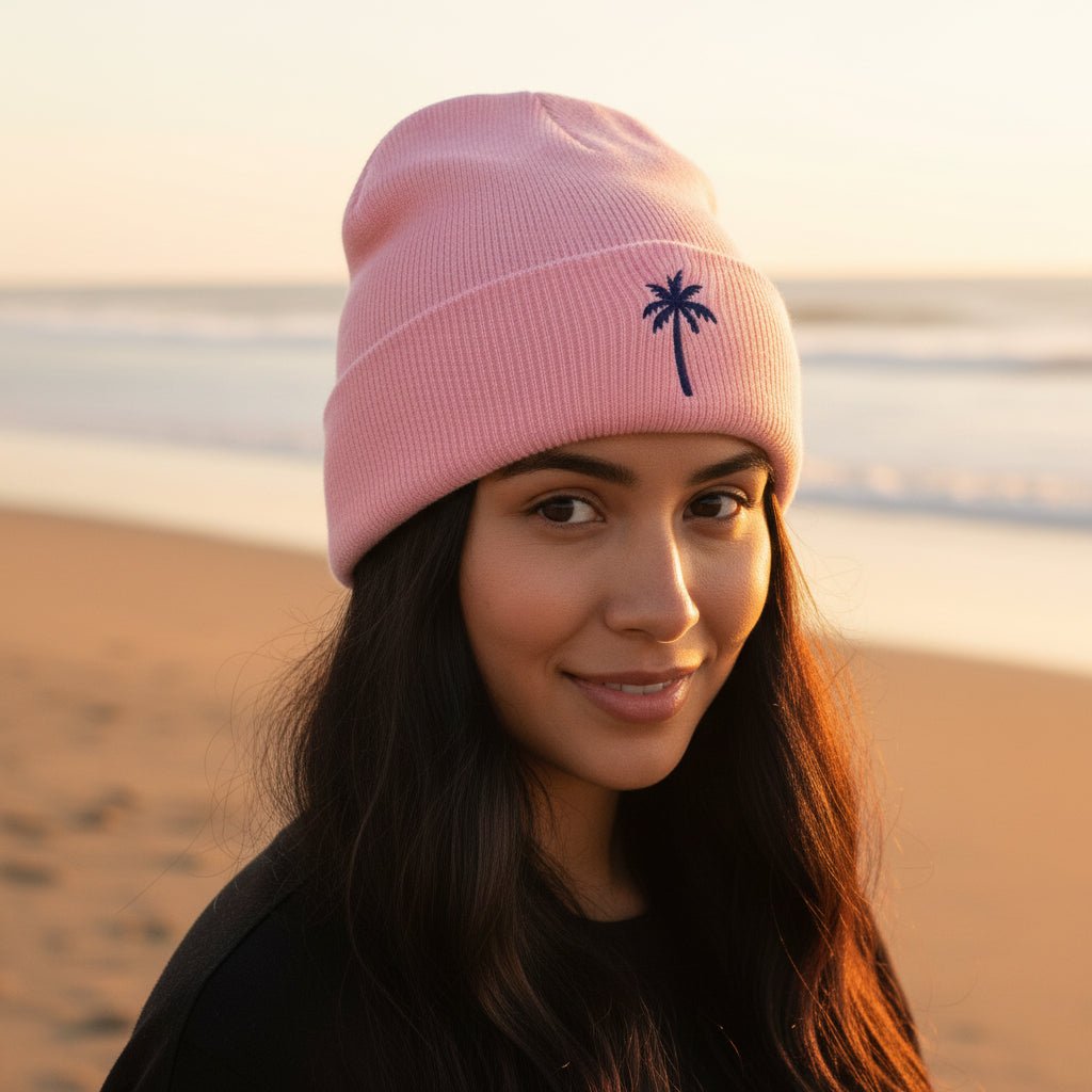 A light pink knit beanie hat with a folded cuff featuring a small dark navy palm tree graphic, shown on a beautiful young lady on Southampton Beach