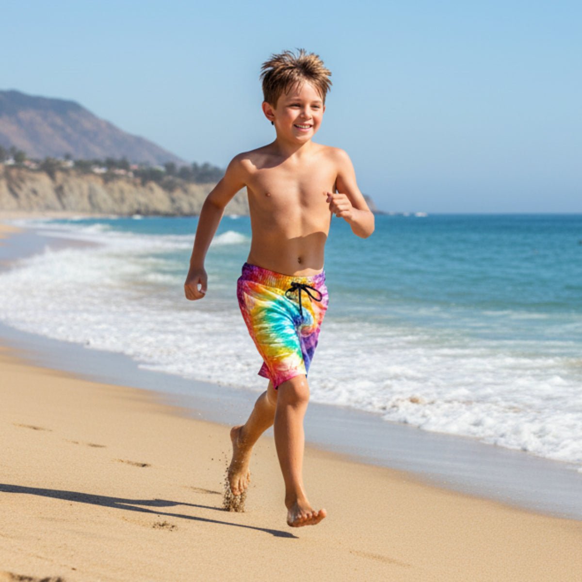Child running on a beach wearing colorful Kids Tie Dye Beach Shorts with ocean and mountains in the background