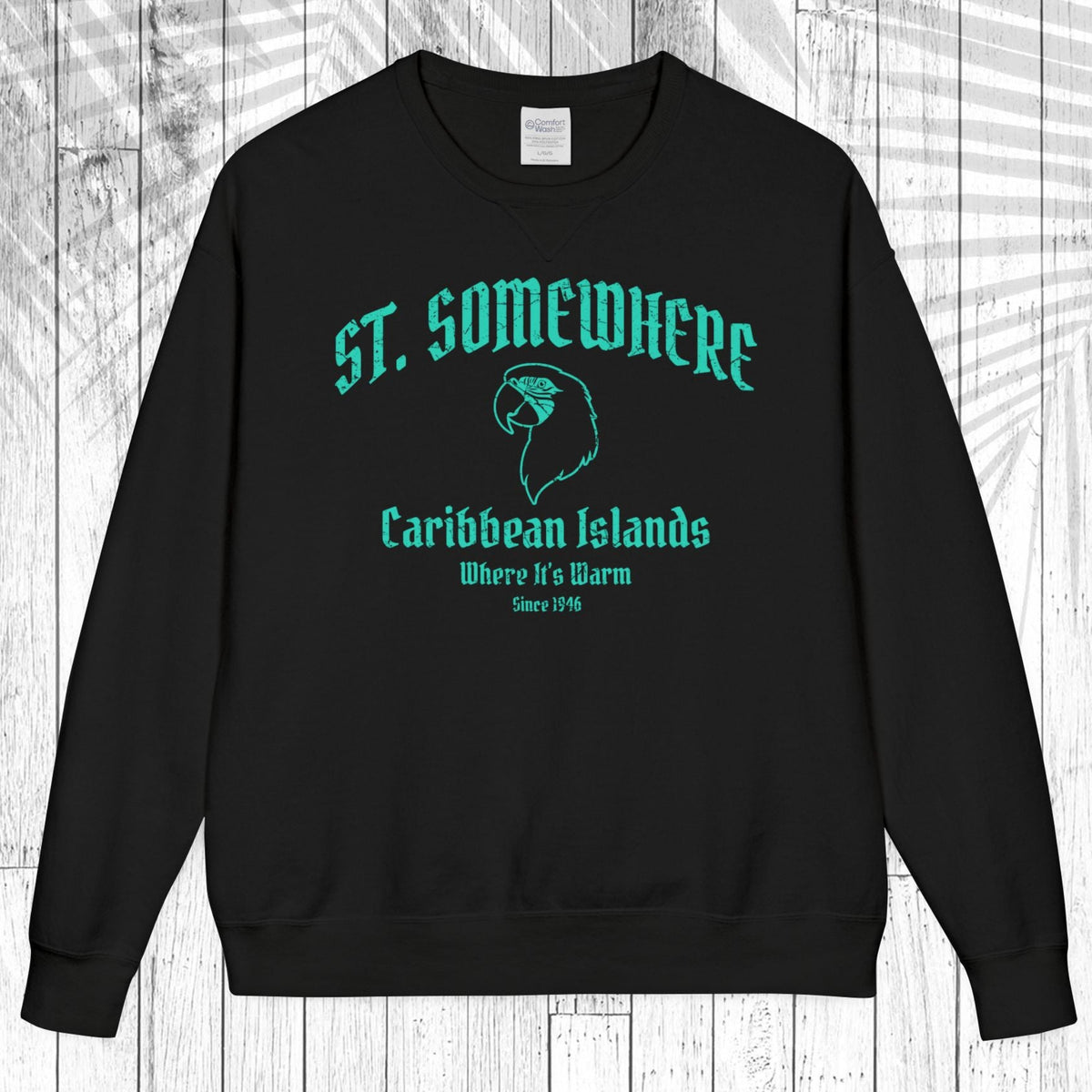 A black crewneck sweatshirt with a teal graphic print on the front reading 'ST. SOMEWHERE Caribbean Islands Where It's Warm Since 1994' with a bird silhouette, displayed flat against a white wood plank background.