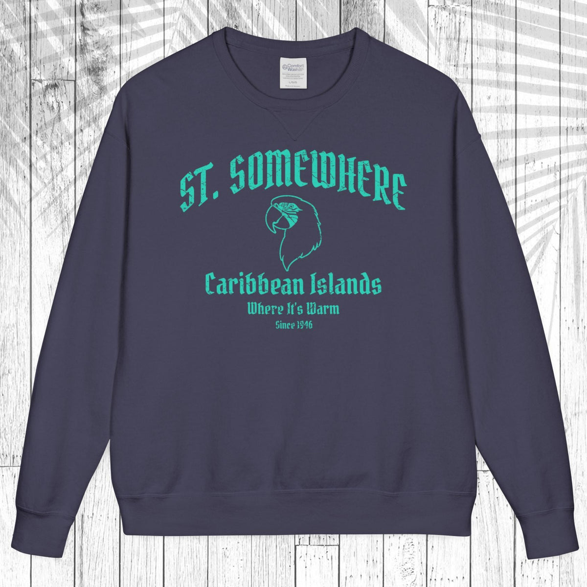 A navy blue crewneck sweatshirt with a teal graphic print reading 'ST. SOMEWHERE Caribbean Islands Where It's Warm Since 1994' and a bird silhouette, displayed flat against a white wood plank background.