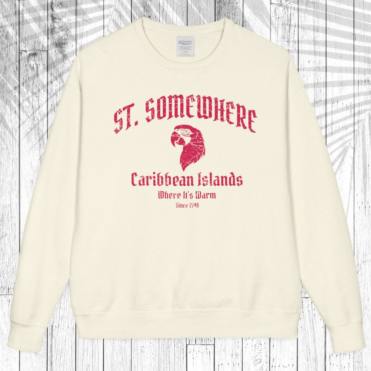 An off-white crewneck sweatshirt with a red graphic print reading 'ST. SOMEWHERE Caribbean Islands Where It's Warm Since 1946' with an eagle head illustration, displayed flat against a white wood plank background.