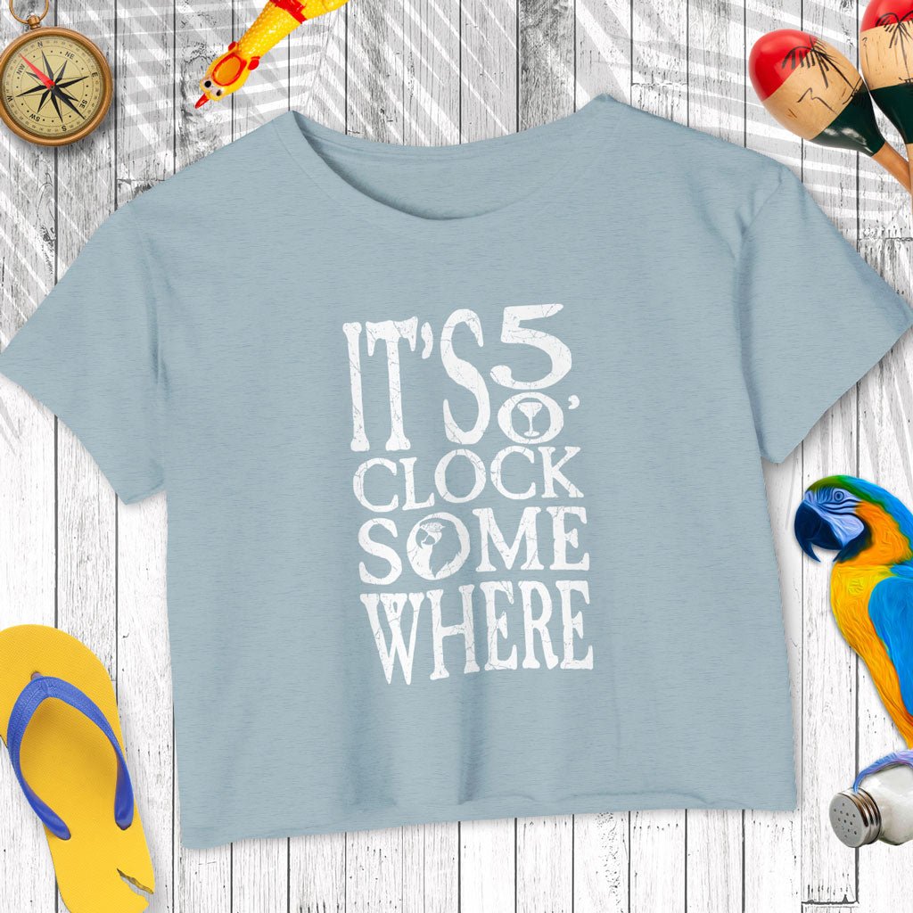 Light blue t-shirt with 'It's 5 o'clock somewhere' text on a wooden surface with decorative items.