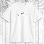 A white short-sleeve crewneck t-shirt featuring a teal graphic of a dog standing on a surfboard on the chest, displayed flat against a white wood plank background with a palm leaf overlay.