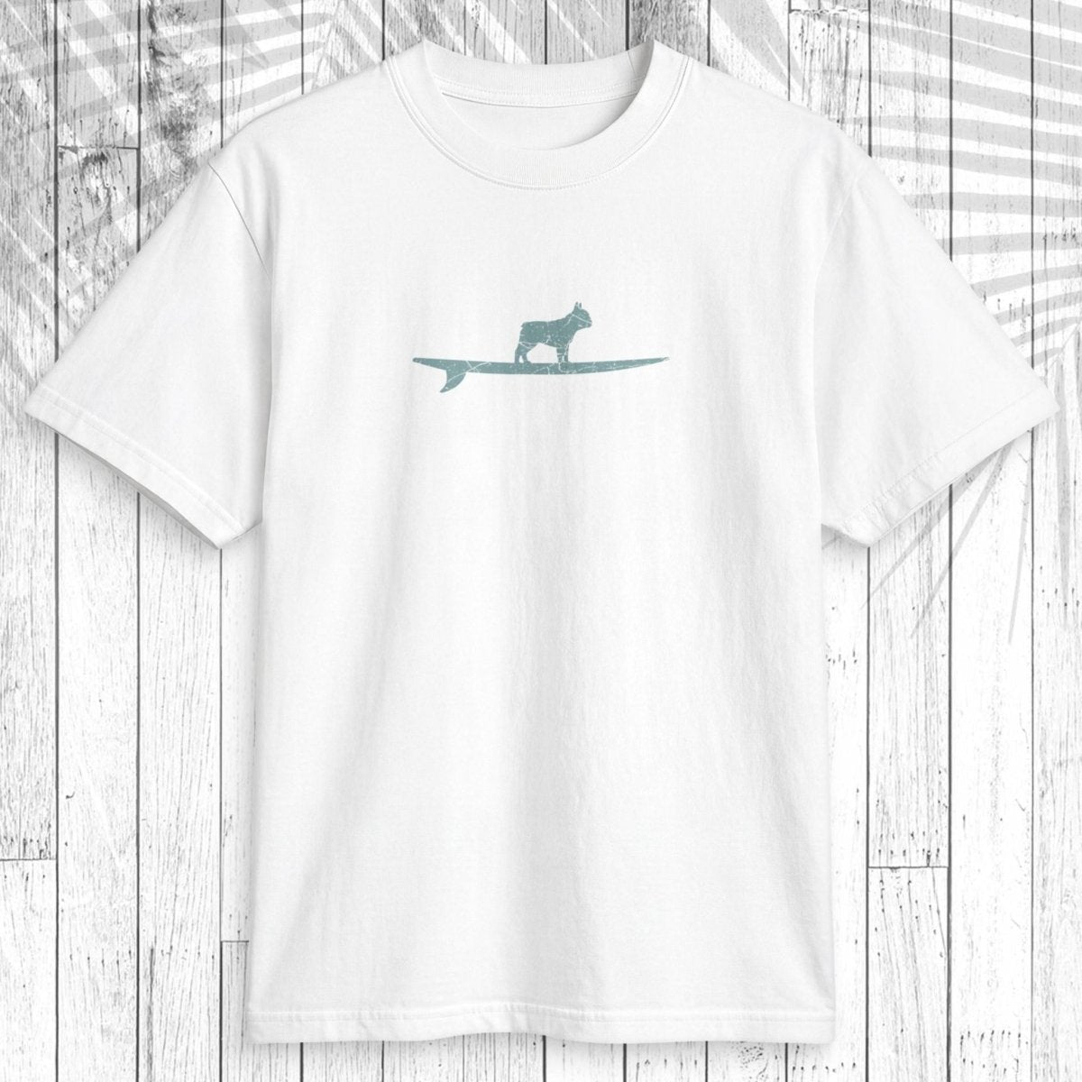 A white short-sleeve crewneck t-shirt featuring a teal graphic of a dog standing on a surfboard on the chest, displayed flat against a white wood plank background with a palm leaf overlay.