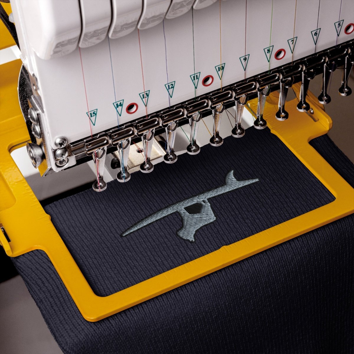Close-up of an embroidery machine stitching a shark design onto dark navy fabric, with the yellow frame and machine components clearly visible.