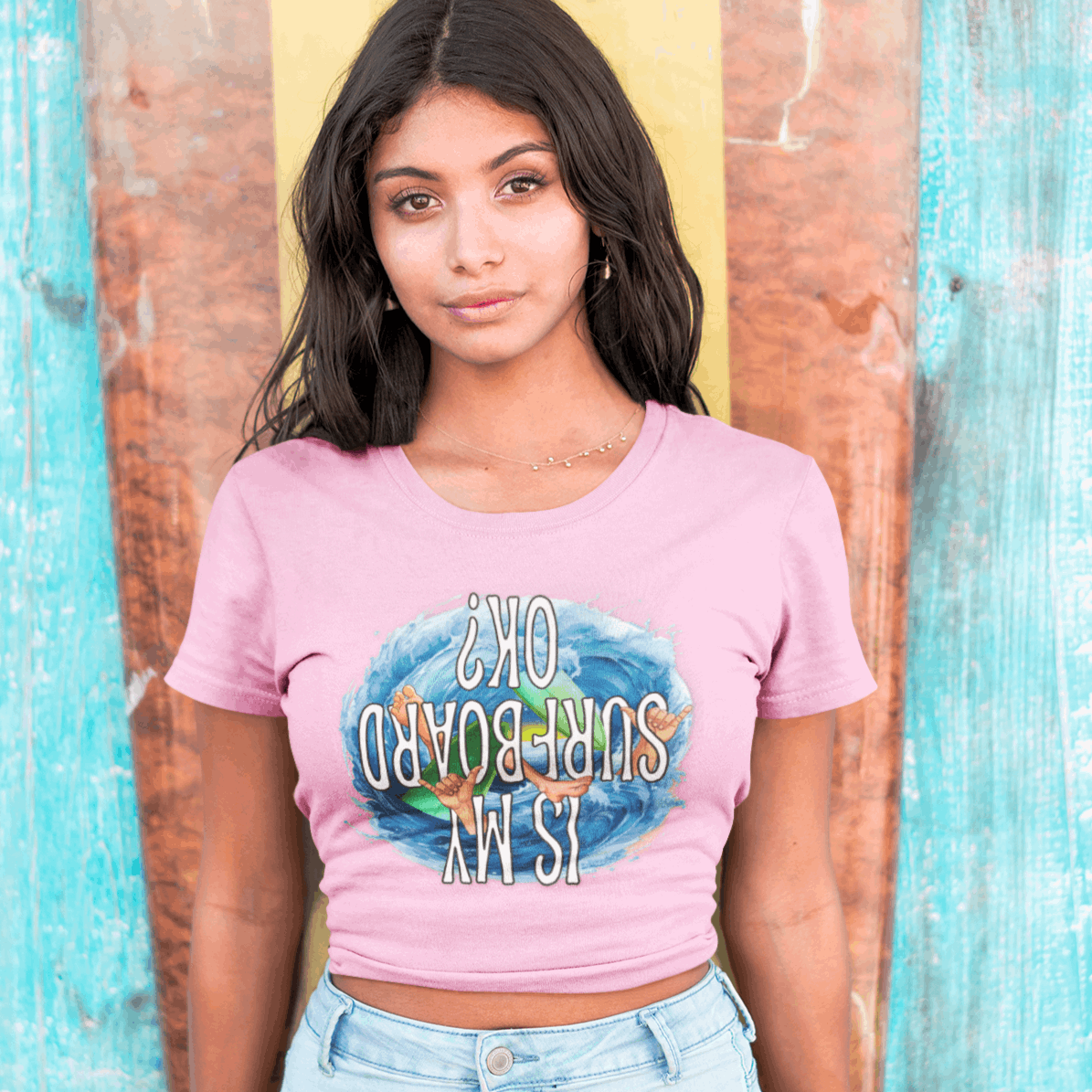 Surfer girl leaning against a surfboard wearing a bubble gum t-shirt from Bella Canvas with funny surfer graphic. 