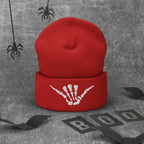 A red knit beanie with a white skeleton hand making Shaka Sign graphic on the cuff, displayed against a textured gray background with Halloween-themed props including spiderwebs, bat wings, and 'BOO' letter cutouts.