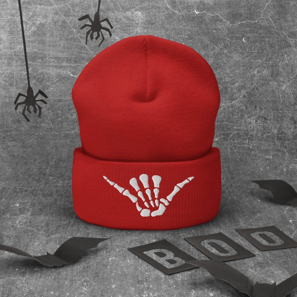 A red knit beanie with a white skeleton hand making Shaka Sign graphic on the cuff, displayed against a textured gray background with Halloween-themed props including spiderwebs, bat wings, and 'BOO' letter cutouts.