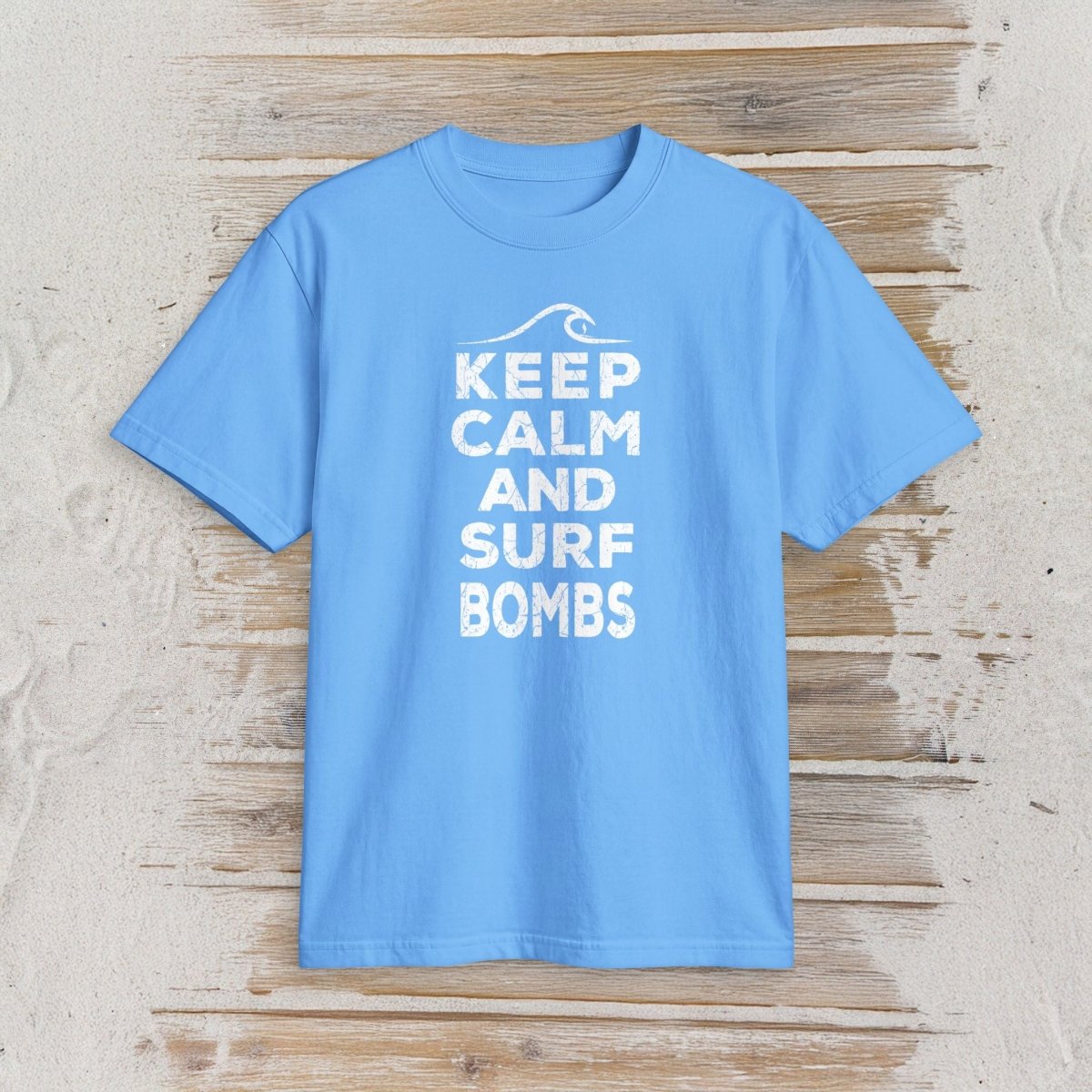 A light blue short-sleeve t-shirt with a white graphic print reading 'KEEP CALM AND SURF BOMBS' and a small wave icon above, displayed flat on a rustic light wood background.