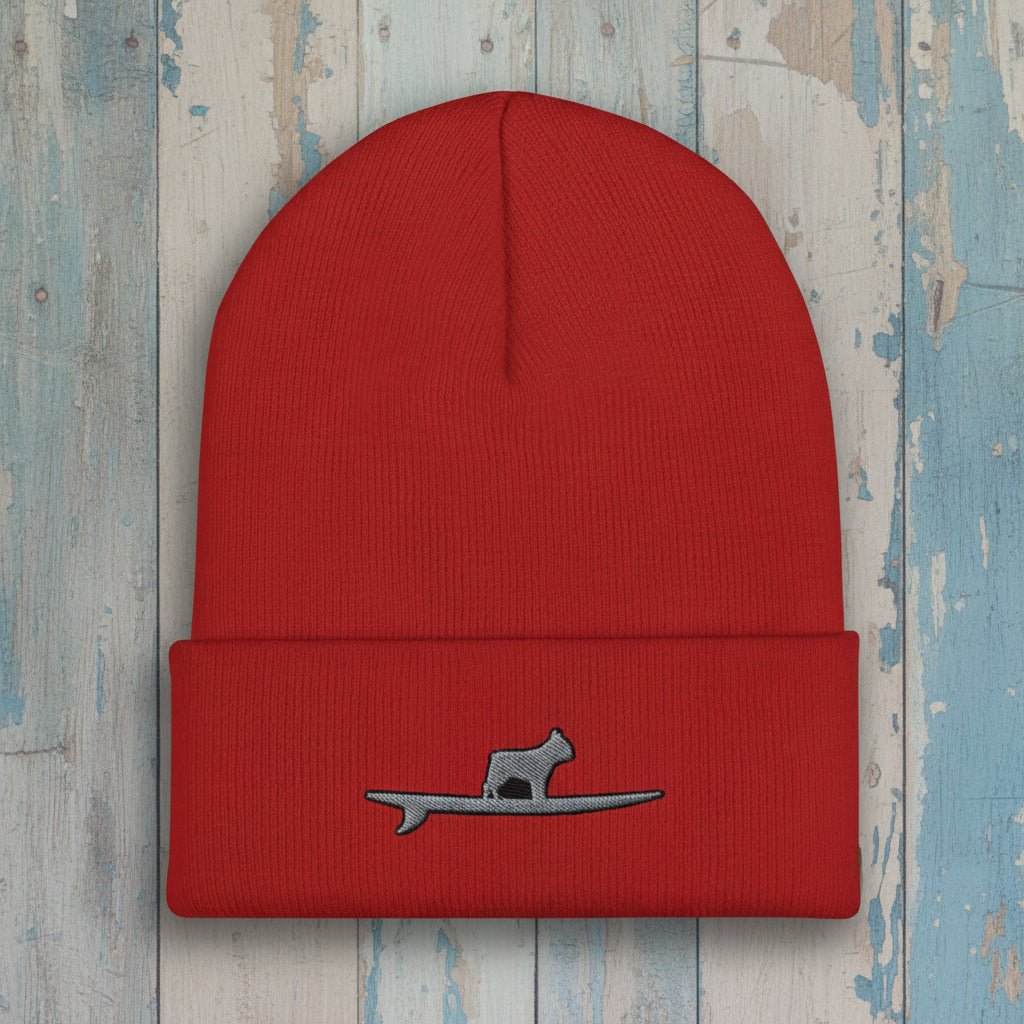 A red ribbed knit beanie with a folded cuff, featuring a small gray embroidered Frenchie silhouette sitting on a surfboard on the front.