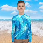 Teen wearing a blue wave patterned rash guard on a beach with ocean and sky in the background