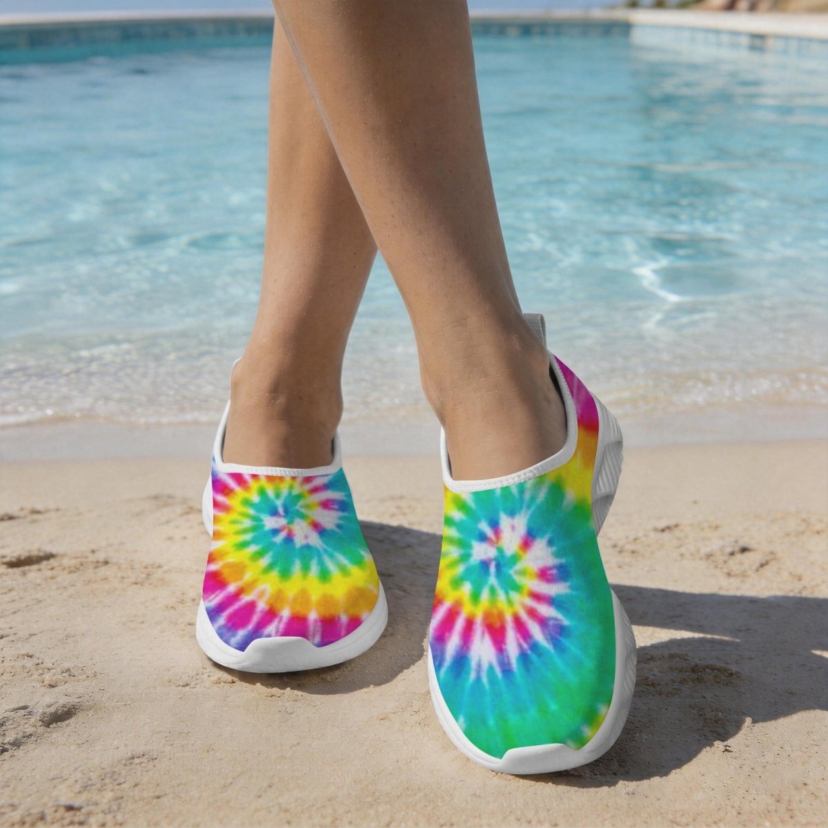 A person standing on a sandy beach by a swimming pool wearing slip-on water shoes with a bright rainbow tie-dye spiral pattern on the uppers and white soles.