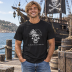 A smiling man wearing a black graphic t-shirt stands on a wooden dock with a pirate ship and sea in the background. The t-shirt features a grayscale graphic of a skull wearing a pirate hat with the text 'You Should Know How To Use A Sword'.