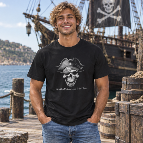 A smiling man wearing a black graphic t-shirt stands on a wooden dock with a pirate ship and sea in the background. The t-shirt features a grayscale graphic of a skull wearing a pirate hat with the text 'You Should Know How To Use A Sword'.