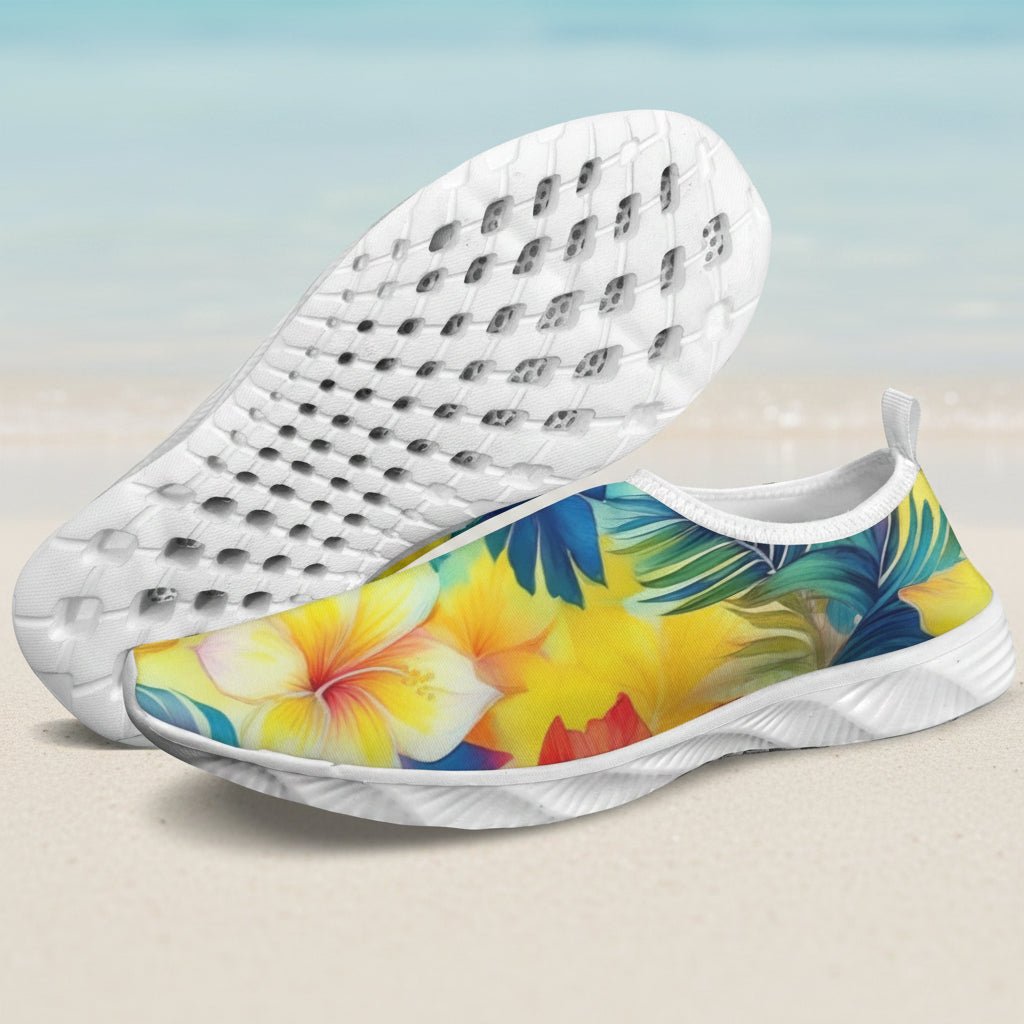 A pair of slip-on water shoes with a white sole and a tropical floral print on the upper. One shoe is shown from the side displaying a yellow hibiscus flower and palm leaves, while the other is turned to show the textured white sole.