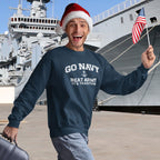 Man wearing a navy sweatshirt with 'Go Navy Beat Army It's Tradition' text, holding an American flag in front of a ship.