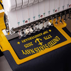 Close-up of an embroidery machine stitching a dark navy fabric with yellow text reading 'United States Navy' and '250th Anniversary 1775 2025' on a yellow guide frame.