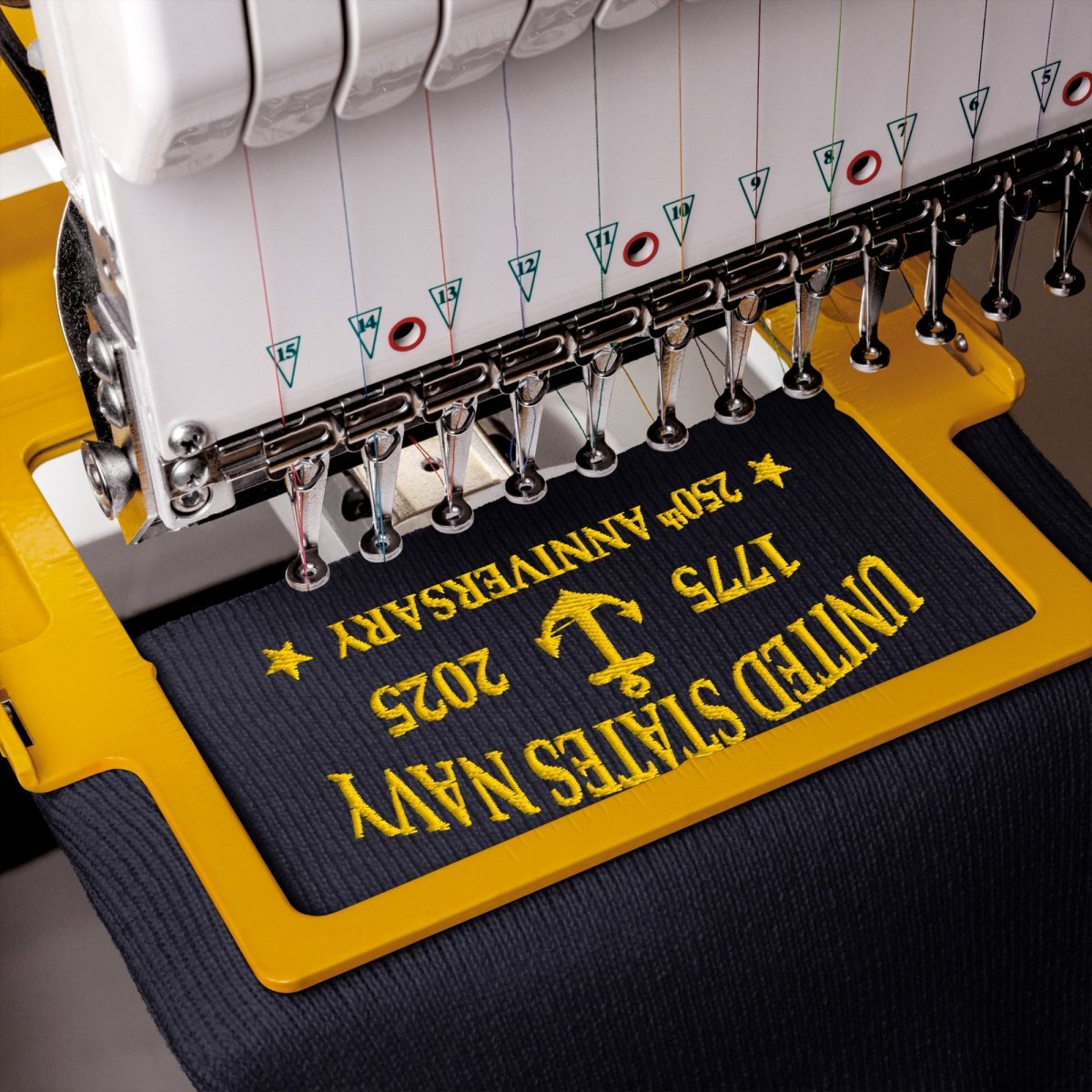 Close-up of an embroidery machine stitching a dark navy fabric with yellow text reading 'United States Navy' and '250th Anniversary 1775 2025' on a yellow guide frame.
