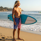 teen surfer dude wearing American Flag board shorts holding surfboard on La Jolla beach with ocean waves in the background