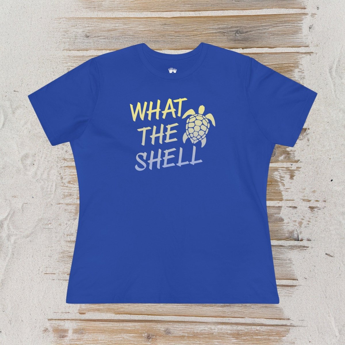 A royal blue short-sleeve t-shirt with a graphic print reading 'WHAT THE SHELL' in yellow and white, accompanied by a yellow sea turtle illustration. The shirt is laid flat on a rustic, light-colored wooden plank background.