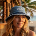 Beautiful blond woman wearing a blue bucket hat with WWJBD embroidered on the front. sitting in a tiki lounge