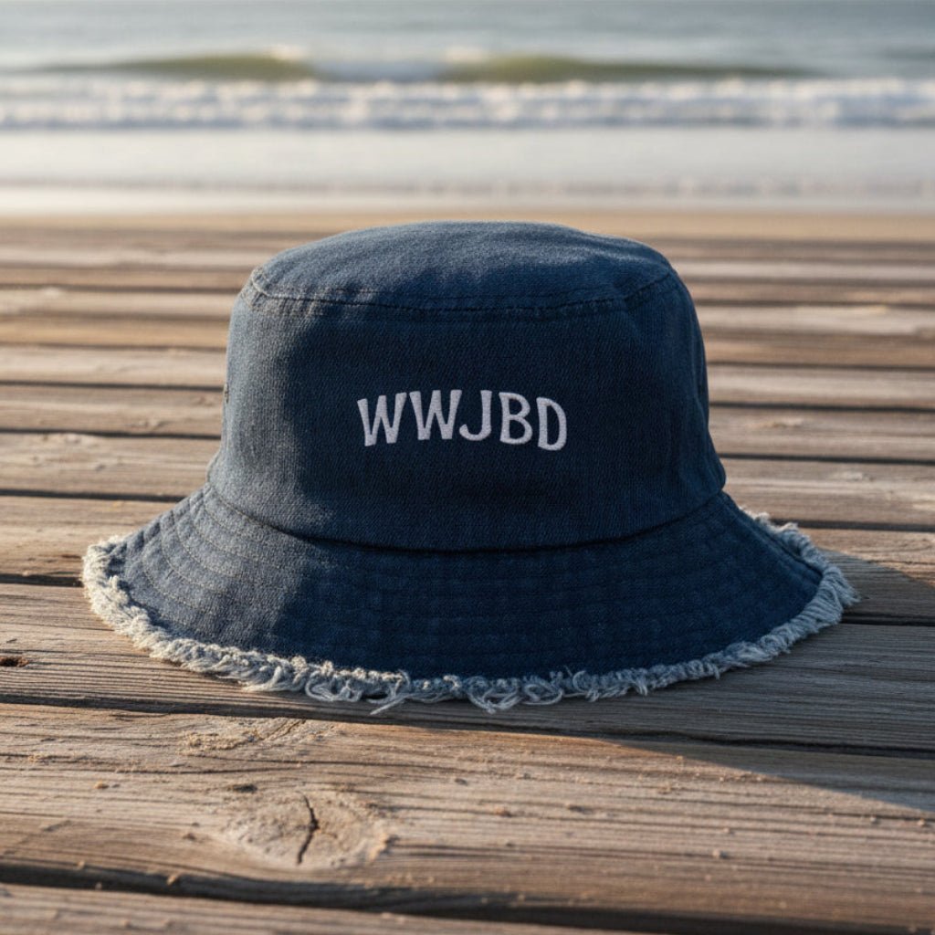 A dark blue denim bucket hat with white embroidered text reading 'WWJBD' sits on a weathered wooden boardwalk with the ocean in the background.