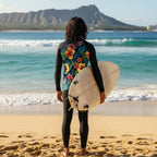 Dude with long black hair looking out at the waves from Waikiki Beach holding a surfboard wearing a long-sleeve shirt with colorful tropical floral design