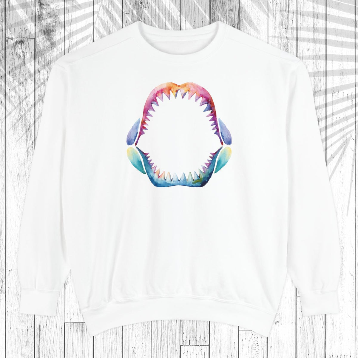 A white crewneck sweatshirt featuring a colorful, watercolor-style graphic of an open shark mouth with sharp teeth on the chest, displayed against a white wood plank background with a faint palm leaf overlay.