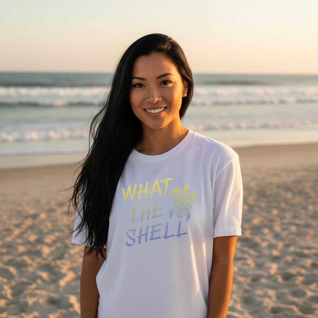 A white short-sleeve crewneck t-shirt hanging on a light wood hanger against a light gray paneled wall. The shirt features a graphic print on the front with the text 'WHAT THE SHELL' in a yellow-to-blue gradient, accompanied by a matching gradient sea turtle illustration.