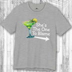 Gray Margarita Party Tee with a graphic of a green cocktail and text 'She's The One To Blame' on a wooden surface