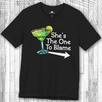 Black Margarita Party Tee with a graphic of a green cocktail and text 'She's The One To Blame' on a wooden surface.