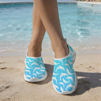 A person standing on a sandy beach by a swimming pool wearing slip-on water shoes with a Blue Dolphin print.