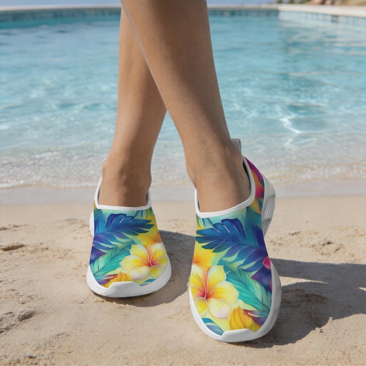 A person standing on a sandy beach with a swimming pool in the background, wearing slip-on Women's Water Shoes with a colorful tropical floral and leaf print.
