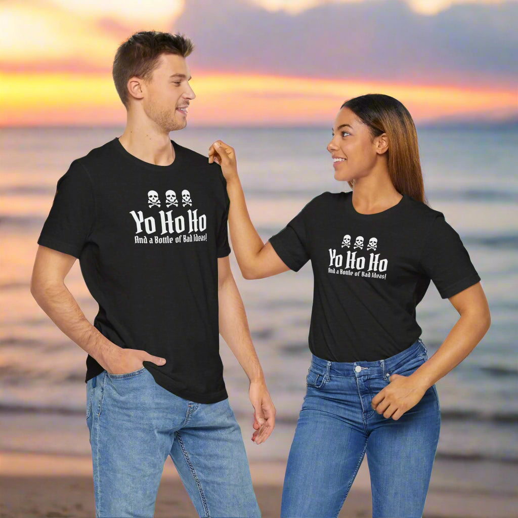 Couple Wearing Bela Canvas Tee Shirts that say "Yo Ho Ho and a bottle of bad ideas" standing on a bech at sunset.