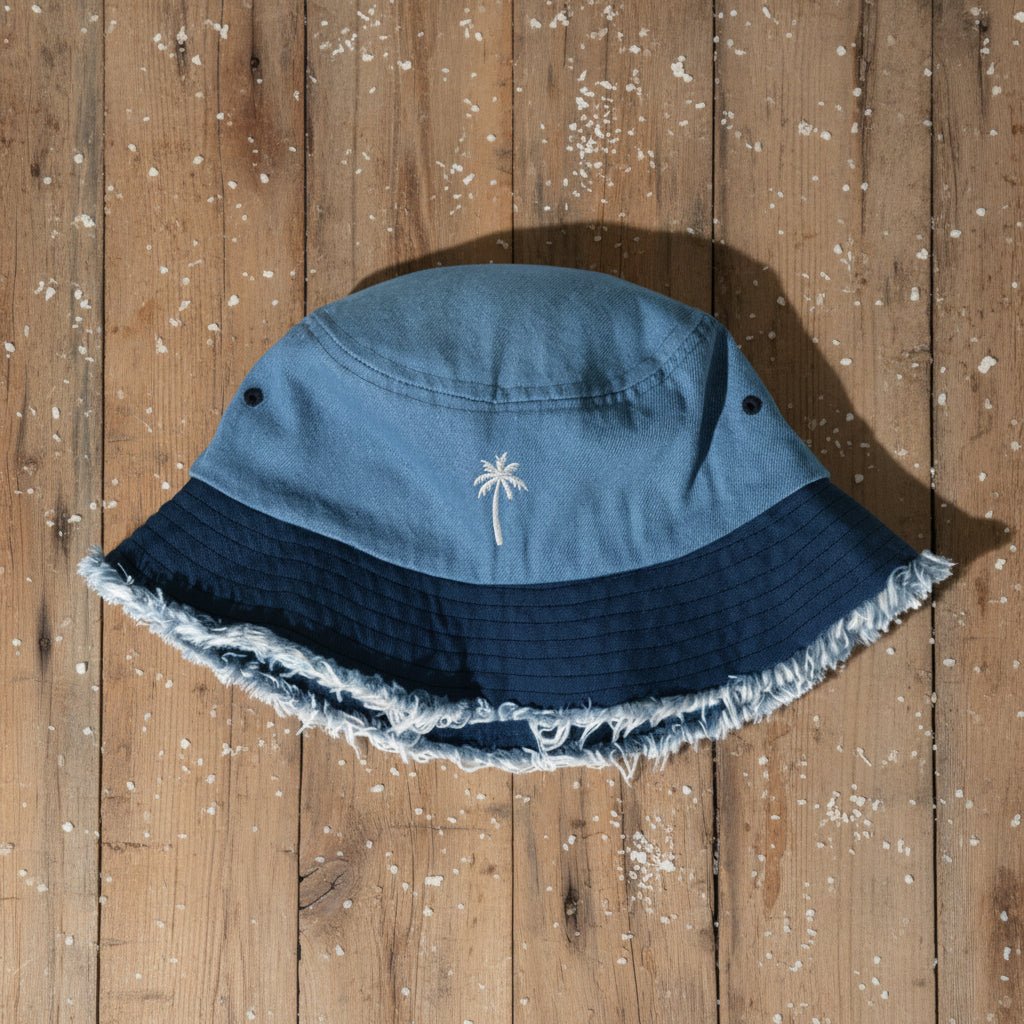 A blue bucket hat with a white palm tree embroidery on the front, resting on a rustic wooden plank background.