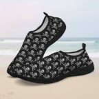 A pair of slip-on water shoes with a black base and an all-over white skull and crossbones pattern, shown on a clean faded beach background.