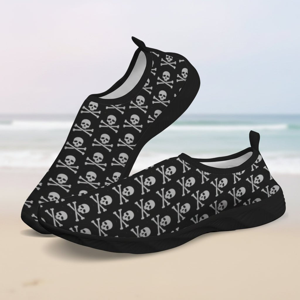 A pair of slip-on water shoes with a black base and an all-over white skull and crossbones pattern, shown on a clean faded beach background.