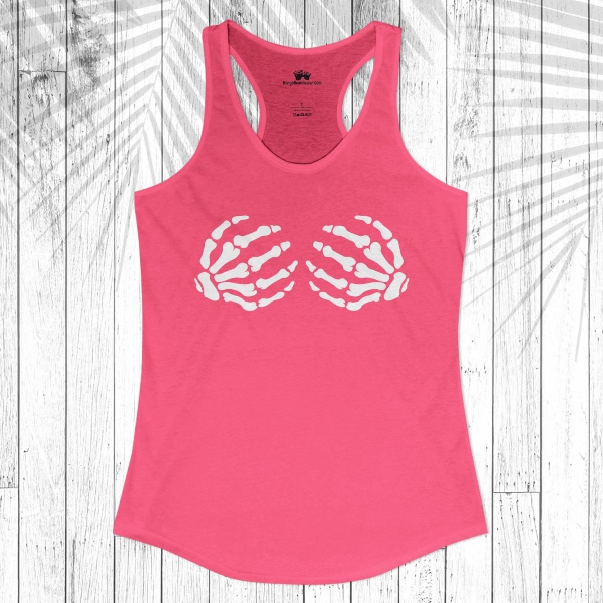 A bright pink racerback tank top with a white graphic of two skeletal hands forming a heart shape on the chest, displayed flat against a white wood plank background with a palm leaf overlay.