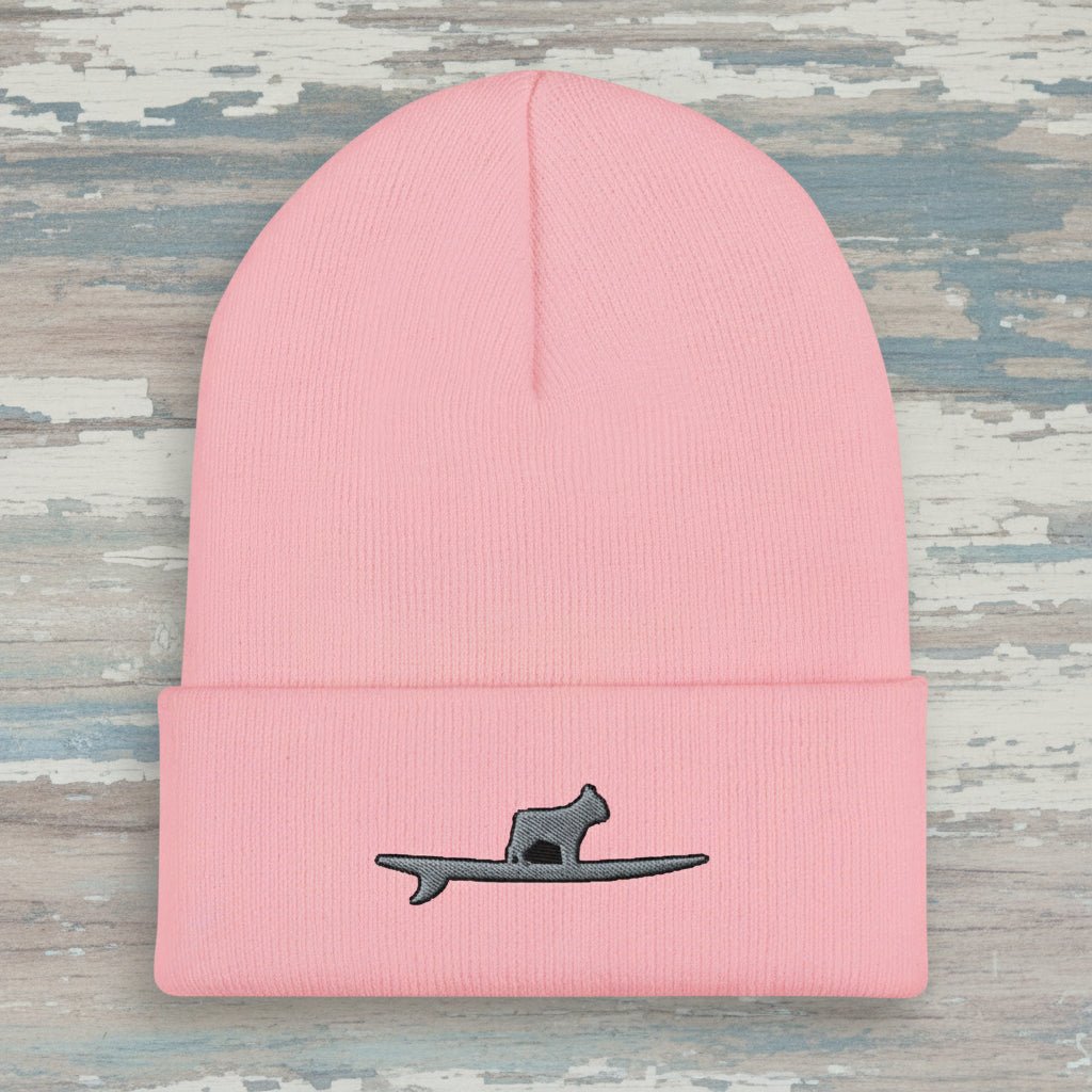 A light pink ribbed knit beanie hat with a folded cuff, featuring a small embroidered design of a gray frenchy standing on a surfboard on the front.