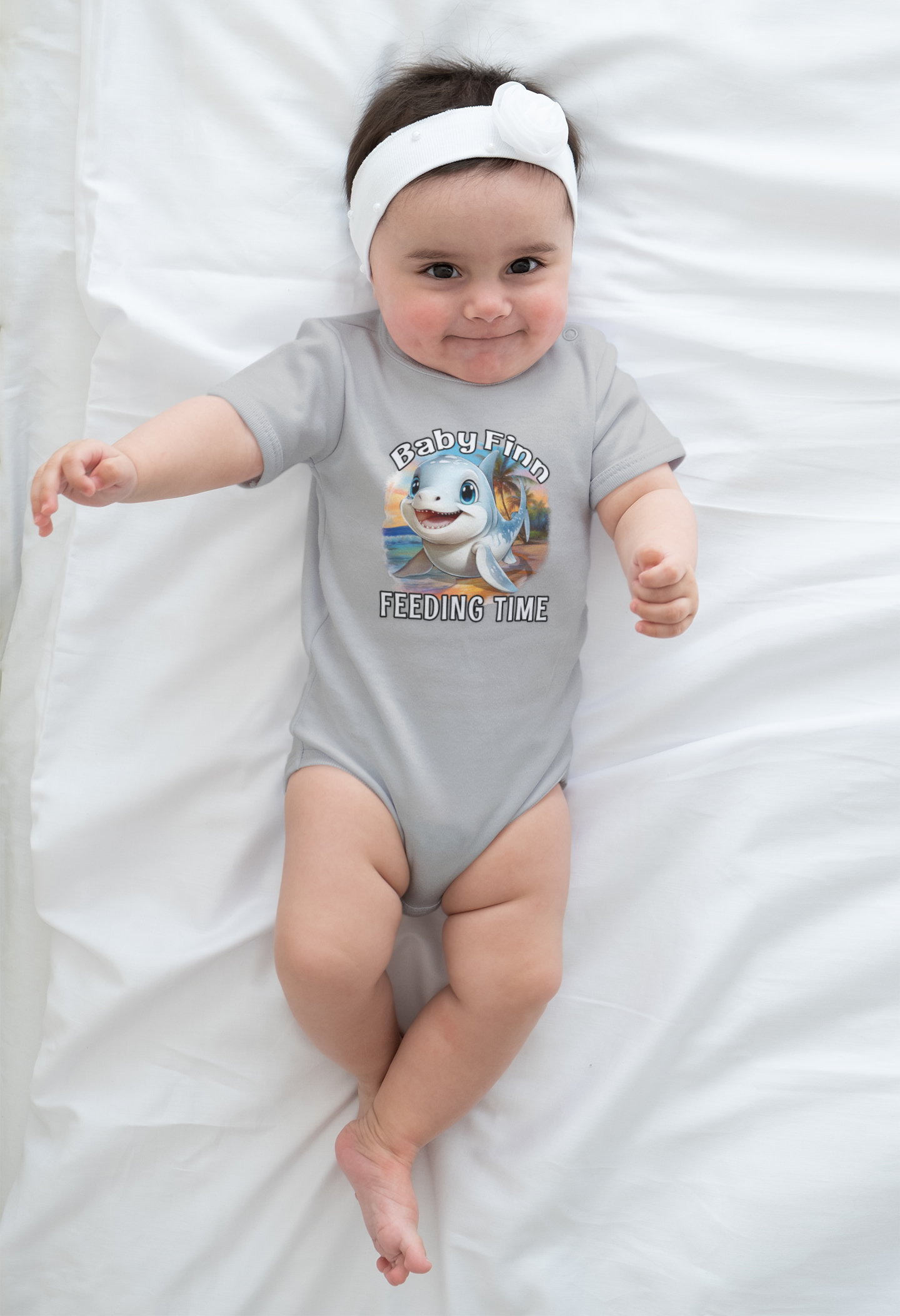 Baby wearing a gray onesie with a cartoon baby shark and text, lying on a white blanket.