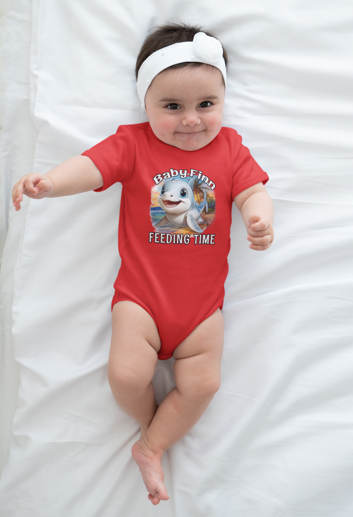 Baby wearing a red onesie with a cartoon baby shark and baby finn text, lying on a white blanket.