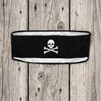A black athletic headband featuring a white skull and crossbones graphic in the center, displayed against a faded nautical wood background