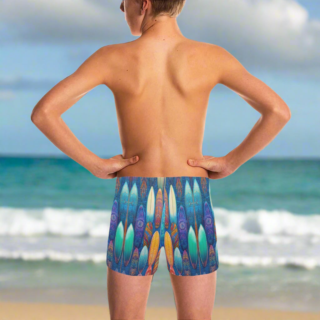 Teen dude wearing colorful surfboard-patterned swim shorts standing on Waikiki Beach looking out over the Pacific